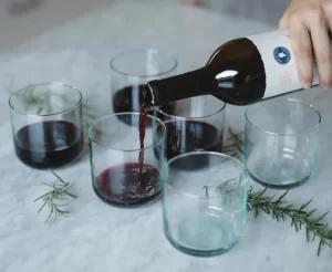 A gray tablecloth with six stemless wineglasses. Someone is pouring red wine from a bottle into the wine glasses. Sprigs of rosemary are placed on the table between the wine glasses as decoration.