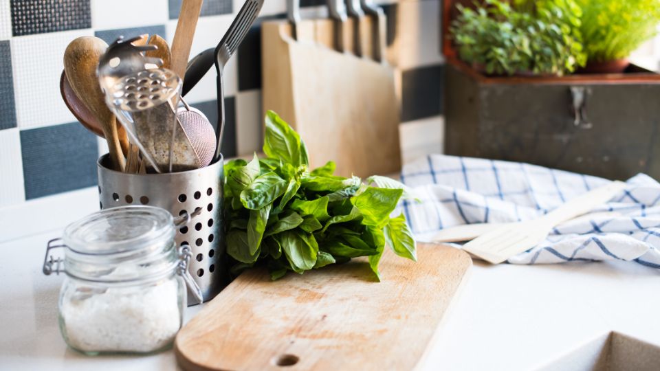 Closeup of a kitchen counter with a wooden cutting board, a metal container filled with kitchen utensils, and a clear glass container with salt. A bunch of fresh basil sits atop the cutting board. In the background are a couple of plants and a blue and white kitchen towel.