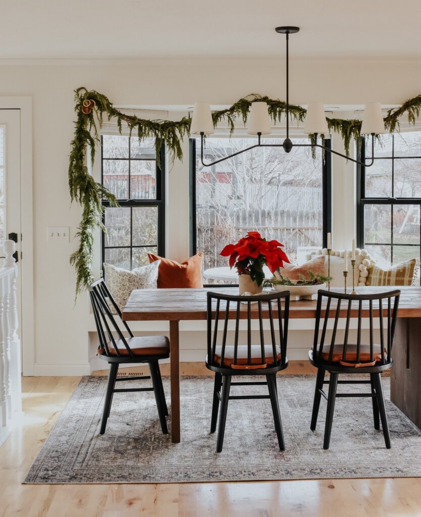Dining room with a table and black windsor chairs. A red poinsettia and three taper candlesticks sit atop the table. An evergreen garland is draped across a bay window with a window seat. 