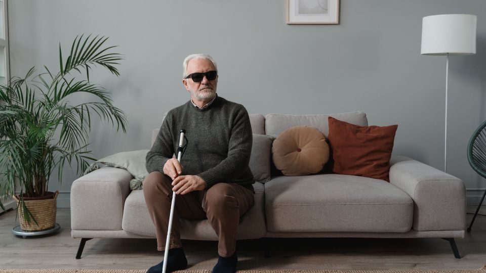 Man with white hair and sunglasses sitting on a tan sofa. He is holding a white cane.