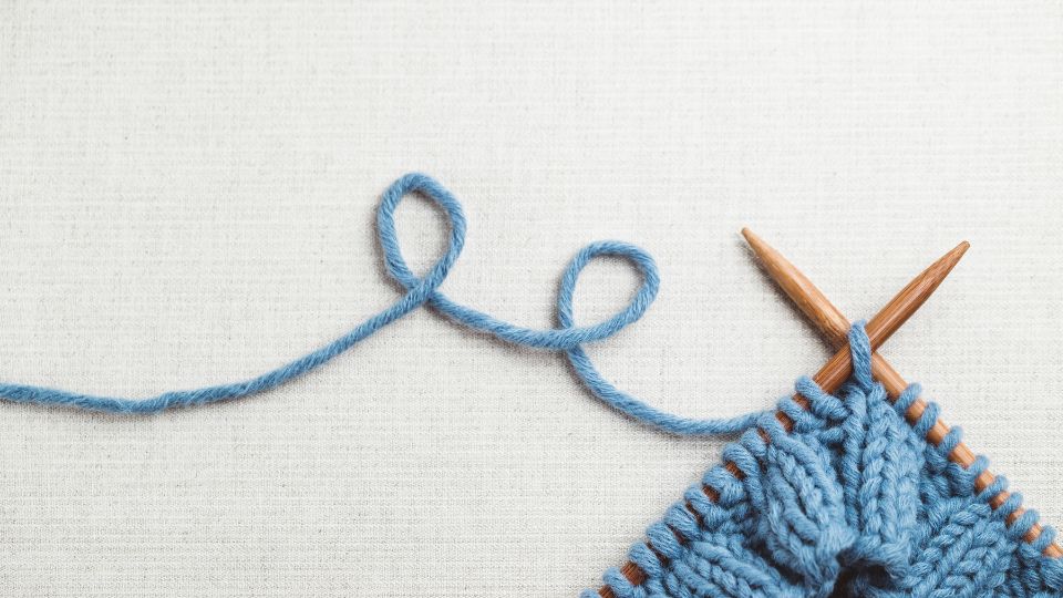 Wooden knitting needles with a project in blue thread. The thread loops off the left side in a playful way.