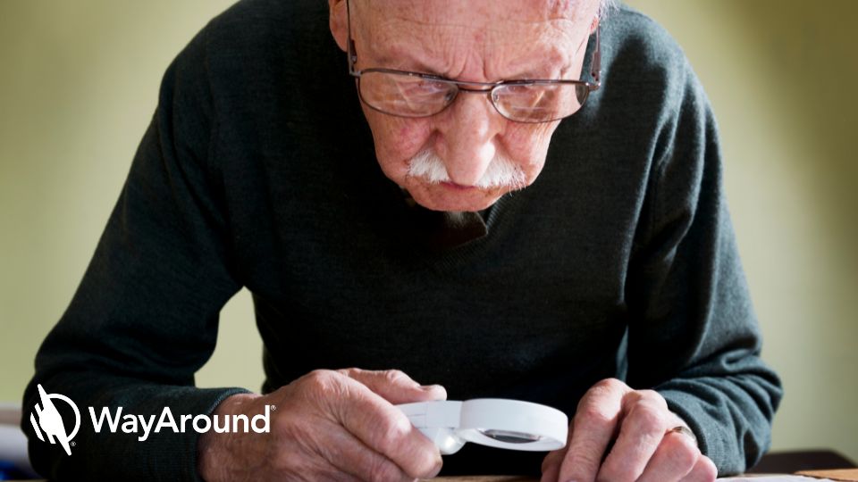 An elderly man with a white mustache and glasses looks down at a table. He is holding a white magnifying glass.