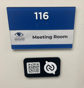 Blue A D A sign that says 116 Meeting room. Beneath the A D A sign is a black sign with a QR code and a white symbol that's a circle with two bumps at the 11 o'clock and 5 o'clock. The white symbol has a black NFC wayfinding symbol printed on top of it.