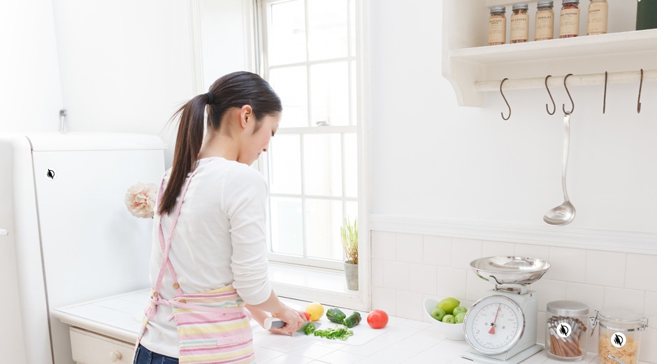 Image of a woman wearing an apron in a kitchen doing meal preparation tasks, chopping vegetables on a cutting board on a countertop with a fridge and canisters of food near her with waytags on them