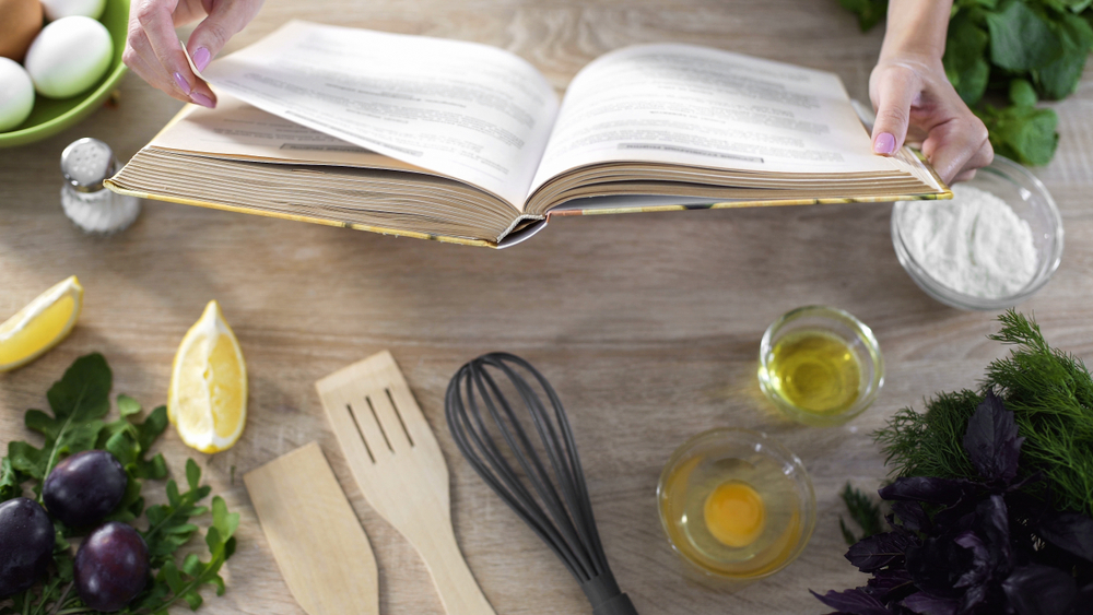 A woman holds a recipe books surrounded by various cooking utensils and basic ingredients such as lemons, eggs, oil, flour, and salt.