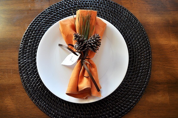 Wooden table with a place setting. There is a dark woven charger, then a white plate. An orange napkin is tied with pinecones and a sprig of greenery. The napkin is on top of the white plate.